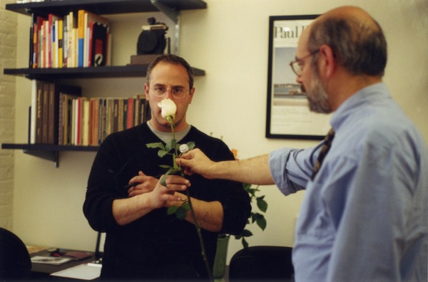 A photo of a man giving another man a white rose while both of them standing in an office.