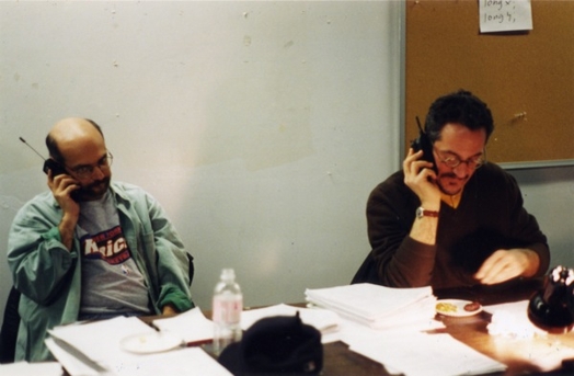 A photo of two people sitting at a table and talking at some old phones with extensible antennas.