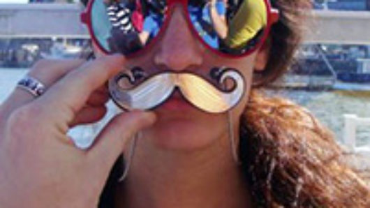 A photo of a woman wearing sunglasses and a false moustache held against her face by another hand.