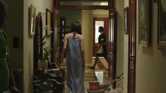 A photo of a woman with a blue dress walking in a hall way while another woman dressed as a maid is walking by.