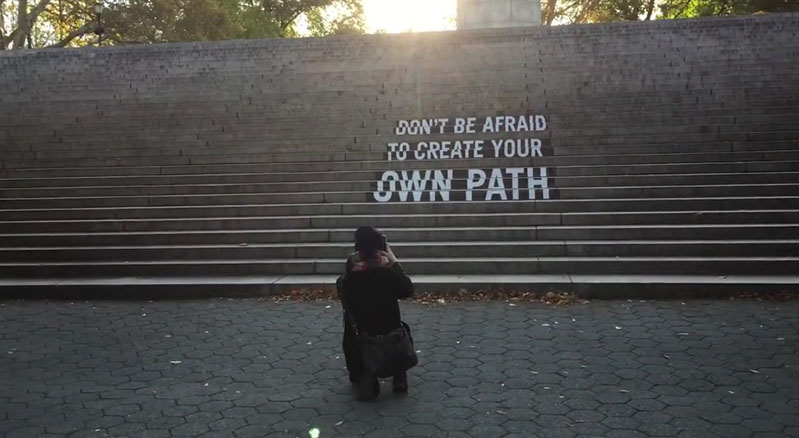 A photo of a person taking a picture of some stairs that have on them a text viewed from a certain angle. The text on the stairs says: Don't be afraid to create your own path. Also above the stairs the sun is shining trough some green trees.