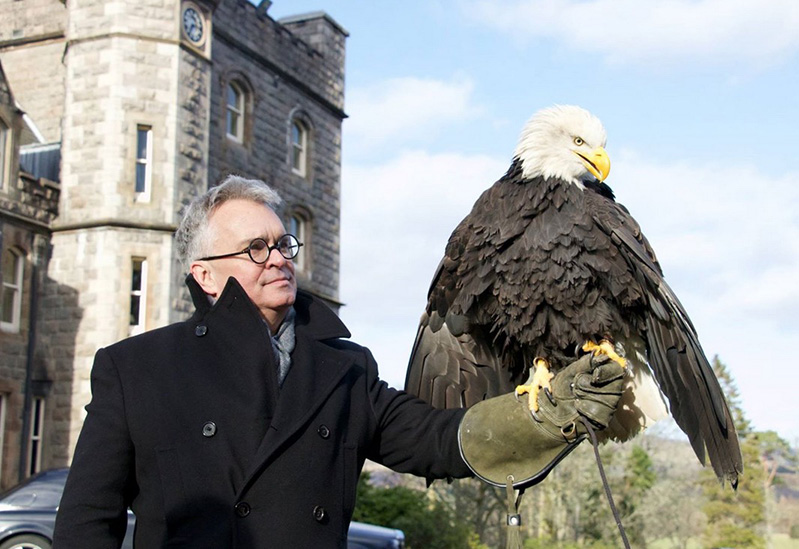A man sitting near an old chateau house while holding in his hand a black and white eagle.