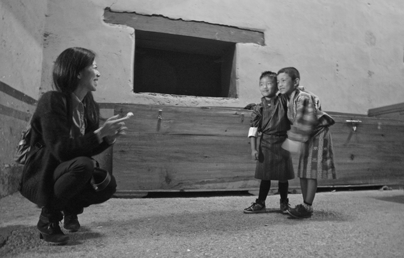 A black and white photo of a woman standing near a building and smiling at two children that hold each other.