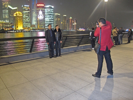 A photo of two people being photographed by another person near a river and behind them a cityscape with color illuminated skyscrapers.
