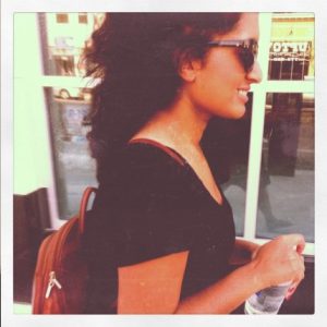 A photo of a woman's profile wearing sunglasses, a backpack and a t-shirt while caring a bottle of water.