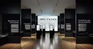 A photo of an art exhibition with black walls and white text on the sides. Also in the middle there are some white posters with some black text that says: SPECTACLES.