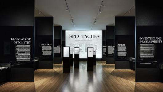 A photo of an art exhibition with black walls and white text on the sides. Also in the middle there are some white posters with some black text that says: SPECTACLES.