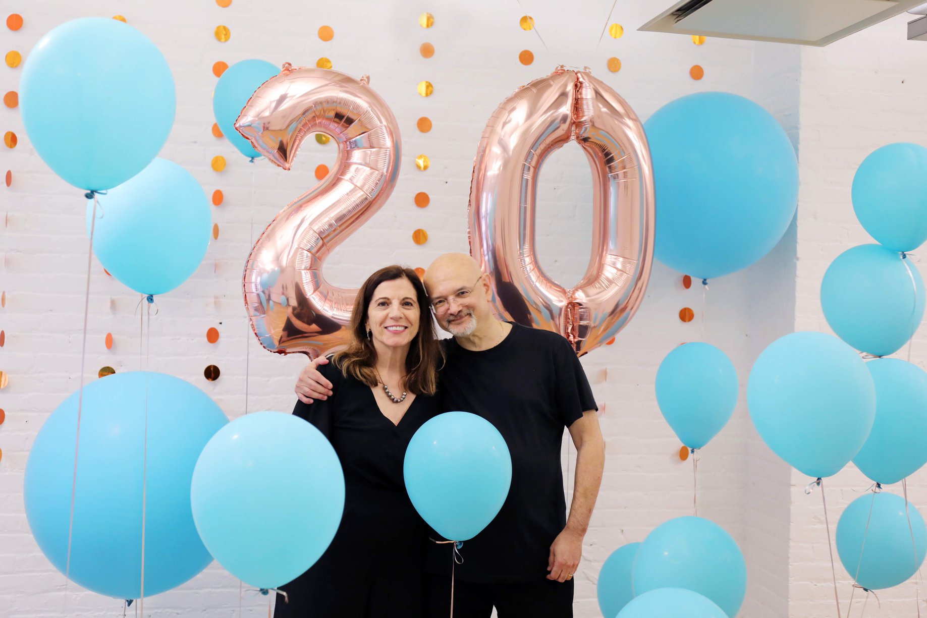 A  photo of a man and a woman surrounded by blue balloons and behind them two balloons that resemble the number twenty.