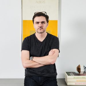 A photo of a man wearing glasses on his head, a watch and a black t shirt sitting in front of a table with stashed books on it. Behind him it's a cream and yellow colored poster.