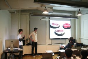 A photo of two people giving a lecture in a classroom while on the screen projector are some 3d pink, black and white logos.