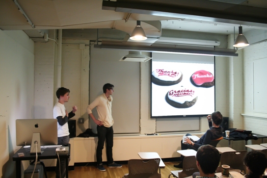 A photo of two people giving a lecture in a classroom while on the screen projector are some 3d pink, black and white logos.