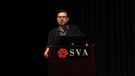 A photo of a man giving a lecture at a stand with SVA logo on it.