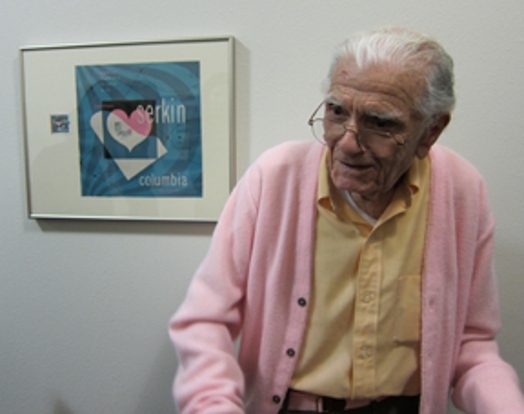 A photo of an old man wearing glasses, a yellow shirt and a pink blouse sitting in front of a blue printing with frame that shows a white and pink heart.