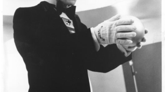 A black and white photo of a man wearing a suit, glasses, a shiny helmet and gloves while holding a ball in his hands.
