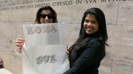 A photo of two women standing near a letter carved stone wall, while holding pieces of paper with text ROMA SVA. The text has ben transferred from the wall engraving to the paper.