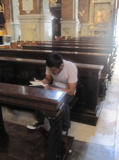 A photo of a man sitting in a church and writing something in a notebook.