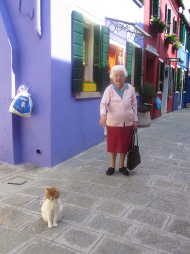 A photo of an old woman and a cat while sitting in a narrow paved street near some colorful houses.