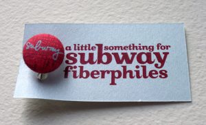 A red button pinned on a piece of white material. The button says subway while on the material the text says: a little something for subway fiberphiles.