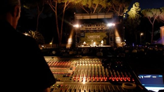 A nightly photo of a sound mixer while in front of it on a distant stage some band is performing.