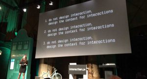 A photo of a person giving a lecture on a stage and showing a text on a projector screen. The text says: - do not design interaction, design the content for interaction.