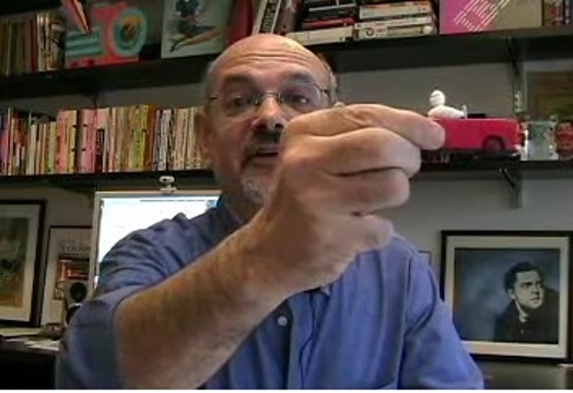 A photo of a man wearing glasses sitting in front of a desk with his hand up and explaining something.