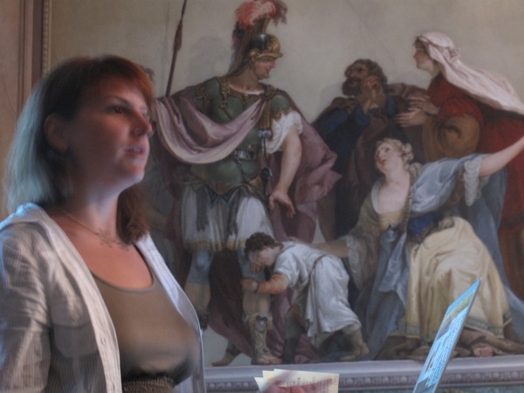 A photo of a woman standing at a laptop near a fresco depicting some people.