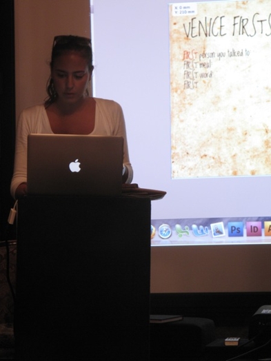 A photo of a woman giving a presentation from a laptop and on the projection screen behind her.
