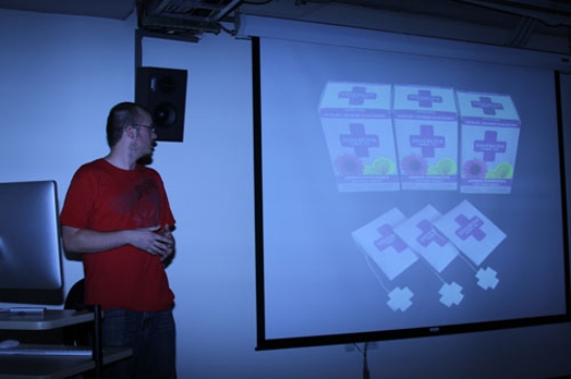 A photo of a man giving a lecture while standing near a screen projector and looking at some tea pack designs.