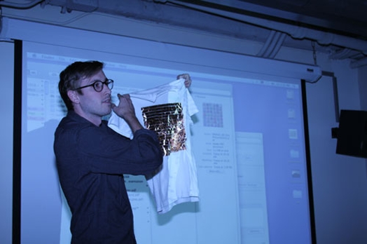 A photo of a man showing a white t-shirt with a golden square to the audience, while standing in front of a screen projector.