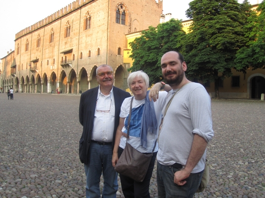 A photo of three people sitting in a paved square near an old building with arches.
