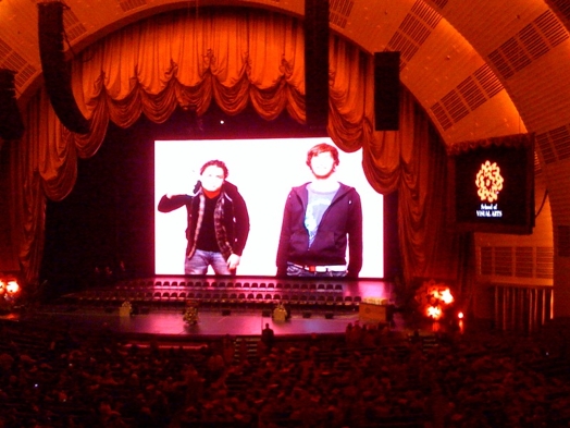 A photo of a semicircular stage with a projected screen in the middle that show a man and a woman talking.