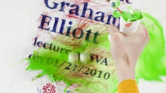 An image of a hand wearing a yellowy blouse, holding a piece of paper and a cup filled with green paint. The paper has blue writings and red lines while green pain is spilled over it.