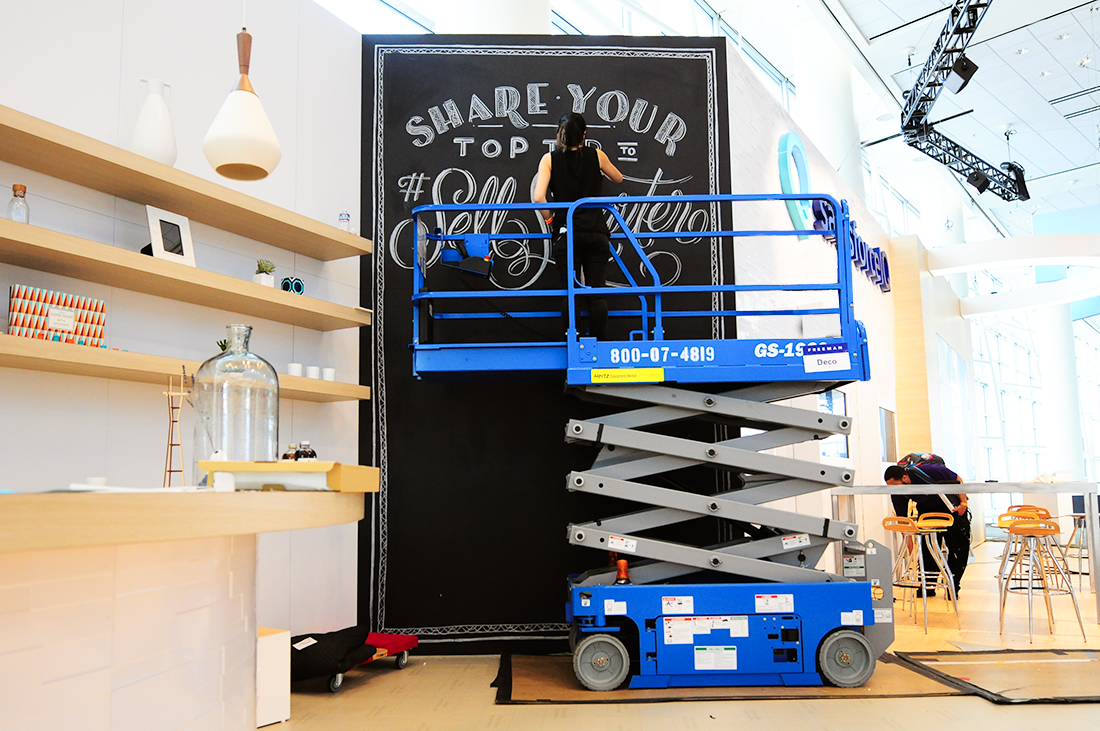 A photo of a person standing in a mobile scissor lift elevator and writing a text on a blackboard that reads: Share Your Top Ten.