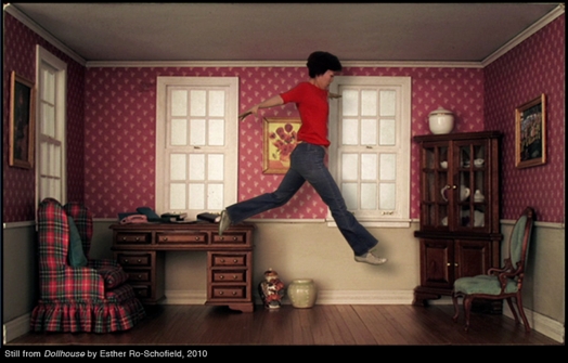 A photo of a woman in red t-shirt jumping mid air in a room with pink wallpaper, an armchair, a green chair, a desk and a cabinet.
