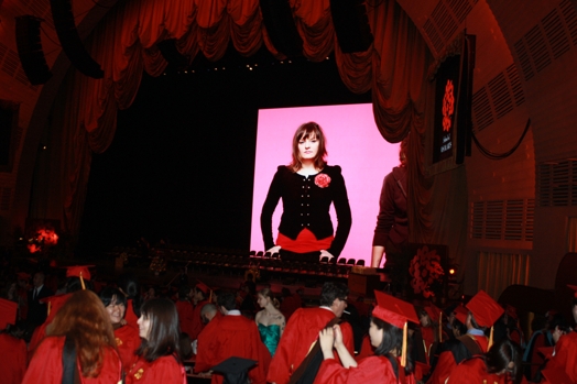 A photo of a theatre stage on which a picture of a girl is projected, while some students talk in the audience.