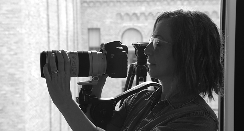 A black and white photo of a woman wearing glasses while holding a DSLR camera in her hands.