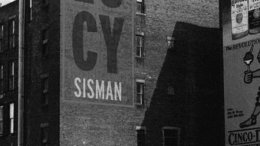A black and white photo of a building and a car near it. On the building is the text LUCY SISMAN.