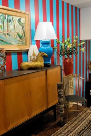 A photo of a room with blue and red stripped wallpaper. A green wood painting, a ceramic horse, a blue lamp, a red vase, a chair and a cabinet are also in the picture.