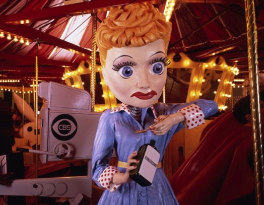A photo of a porcelain woman, wearing a blue dress and sipping from a bottle. Behind it there is what looks like an old filming camera with logo CBS on it and a marry-go-round with yellow lights.