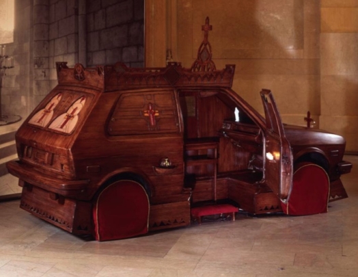 A wood shaped car with open door and red mattresses for wheels.