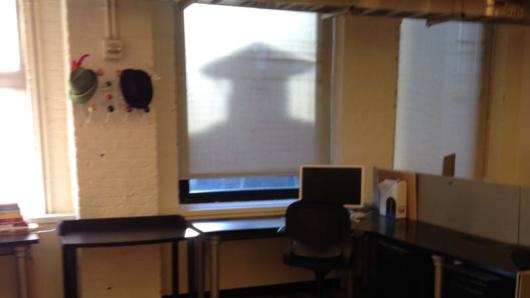 A  photo of a desk wit a pc on it. Behind there is a screen with a shadow of a man wearing a graduation robe and hat.