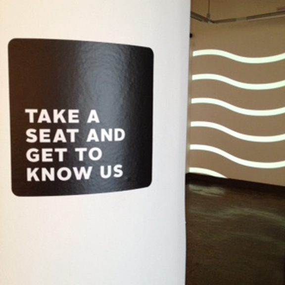 A wall painted with a black rounded square with the shite text: Take a seat and get to know us.