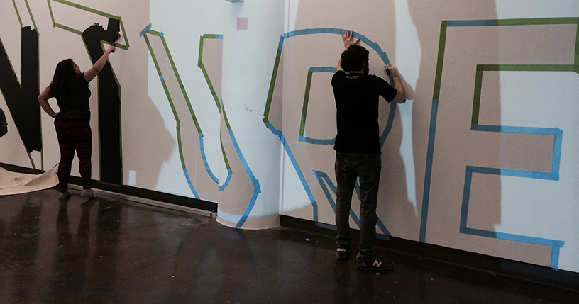 A photo of a man and a woman sticking a blue green duct tape so that letters can be formed and painted between the line. The word VENTURE is being painted.