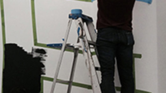 A photo of a man standing on a leader and sticking a blue green duct tape so that letters can be formed and painted between the line.
