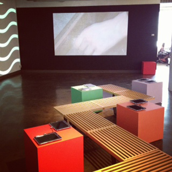 A photo from an art gallery having a wall with wave patterns, some screen projector, some colorful cubical tables and a wood bench between them.
