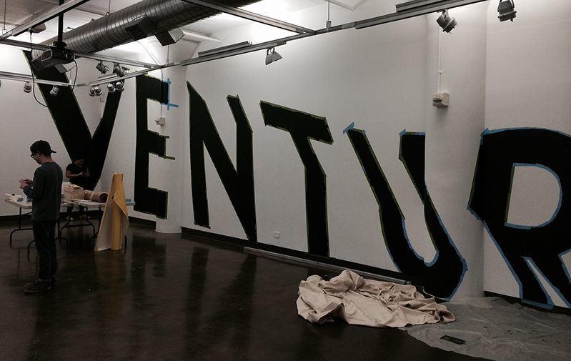 A photo of the word VENTURE painted in black on a white wall in an art exhibition room.