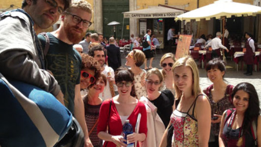 A photo of a group of people in the street while in the background people walk by a yellow building.