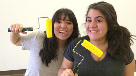 A photo of two girls that are smiling and holding yellow paint rollers.