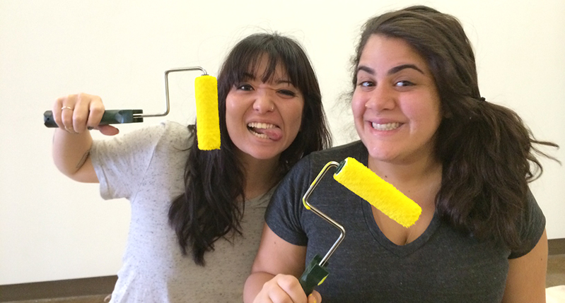 A photo of two girls that are smiling and holding yellow paint rollers.