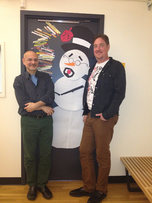 A photo of two people sitting in front of a door with stickers and a snow man with goatee, glasses and hat on it.